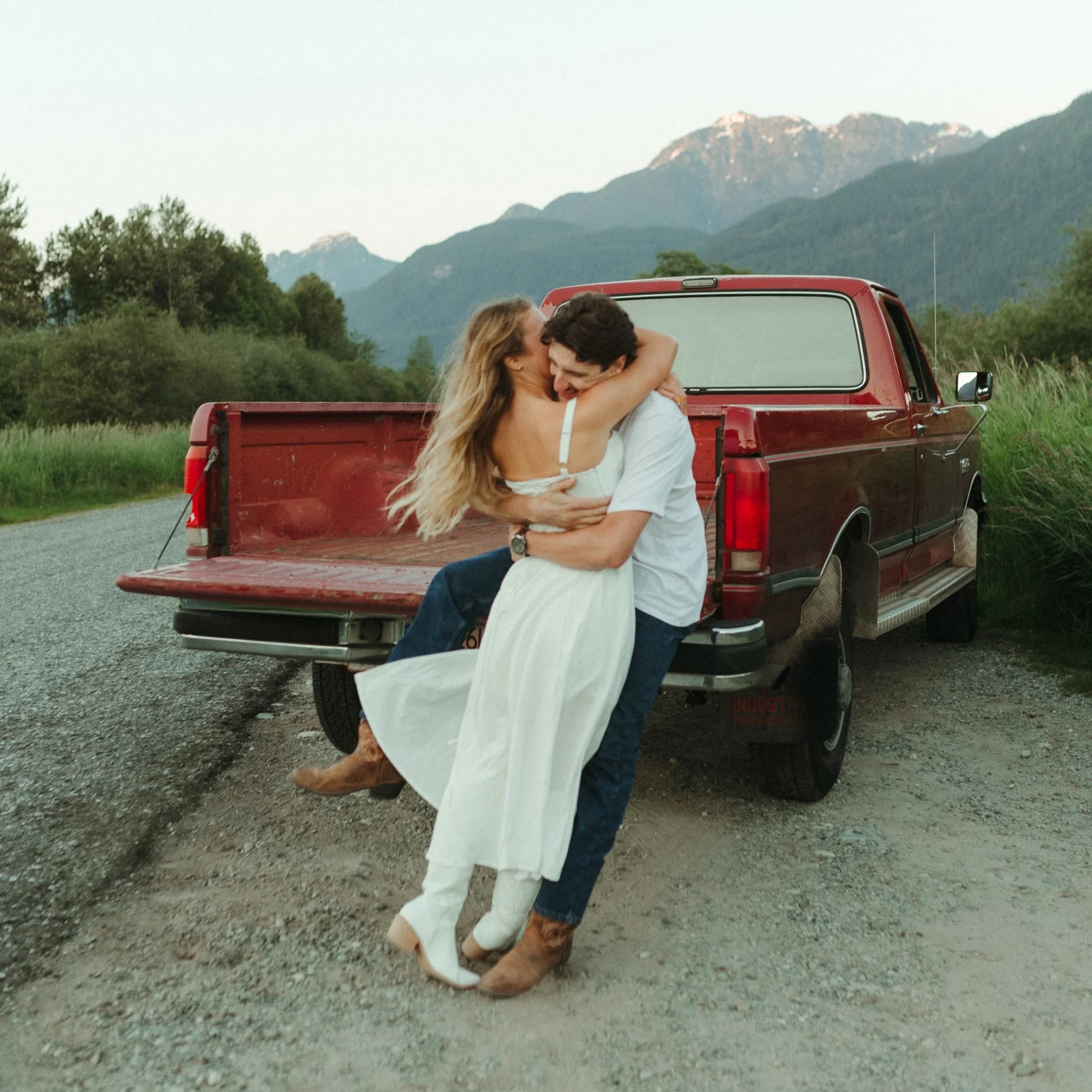 Couple embracing on the back of a red pickup truck, mountains behind.