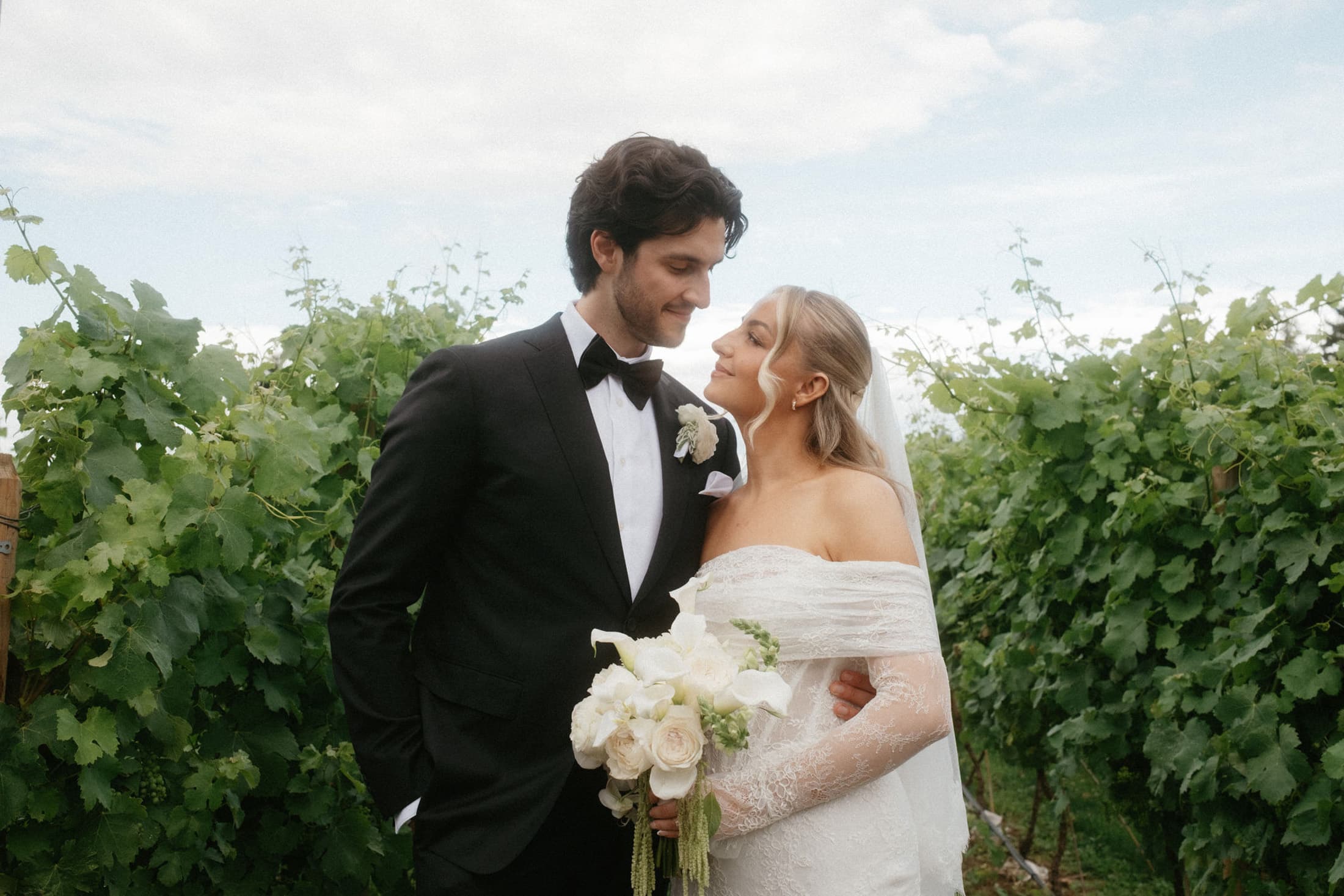 Bride and groom in western-inspired wedding attire standing outdoors beneath a cloudy sky.